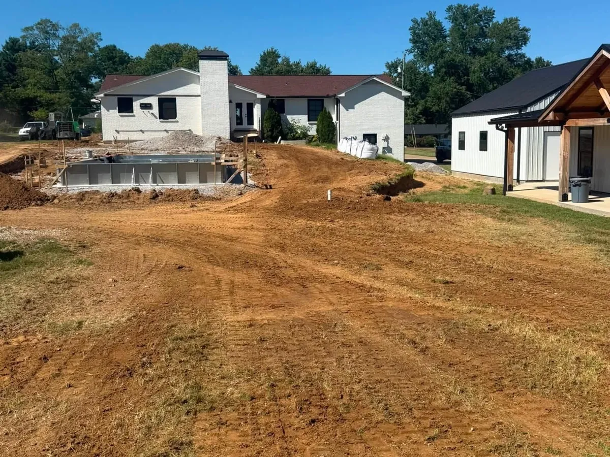 Freshly graded yard beside a white brick house and outbuilding, with an in-ground pool under construction